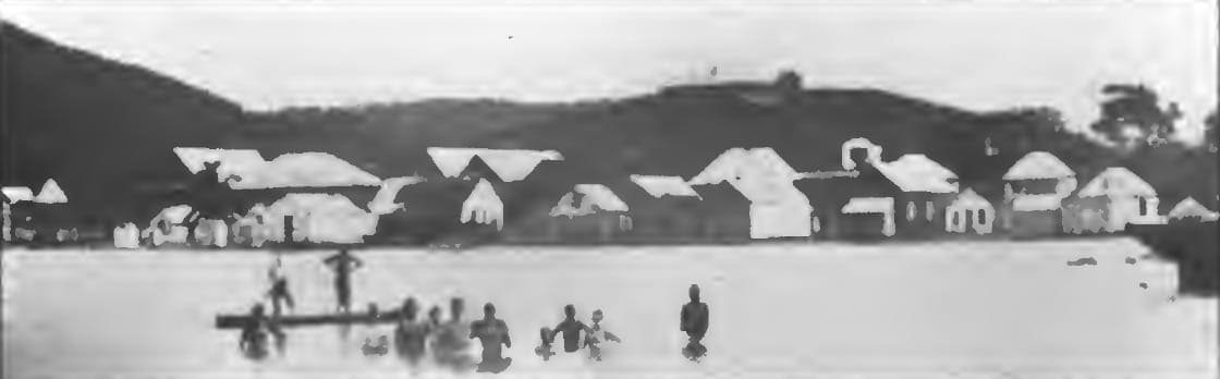 Coxen Hole's waterfront in 1904, with houses along the shore, people wading with a dory, and Government House on the hill behind